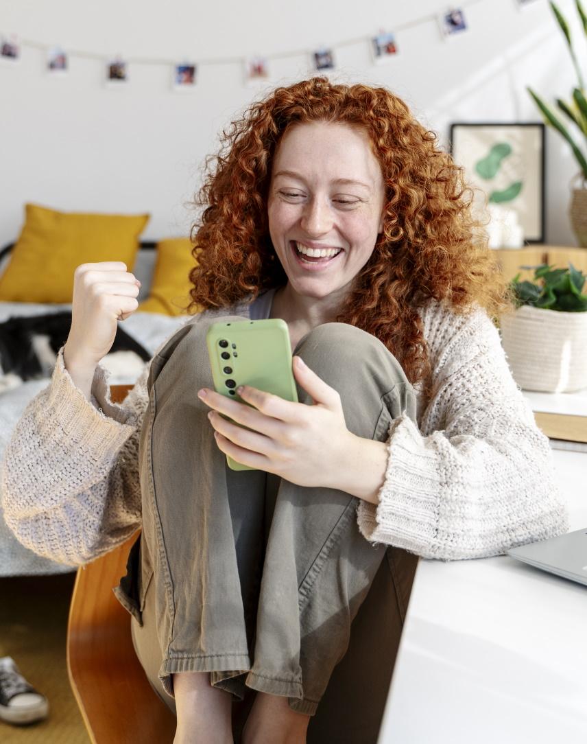 Rothaarige Frau am Schreibtisch schaut auf ihr Smartphone und freut sich