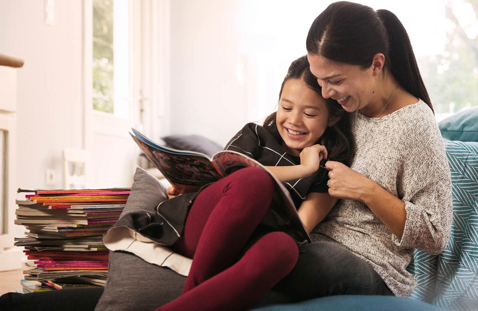 Mutter sitzt mit ihrer Tochter auf der Couch und liest ihr aus Zeitschriften vor