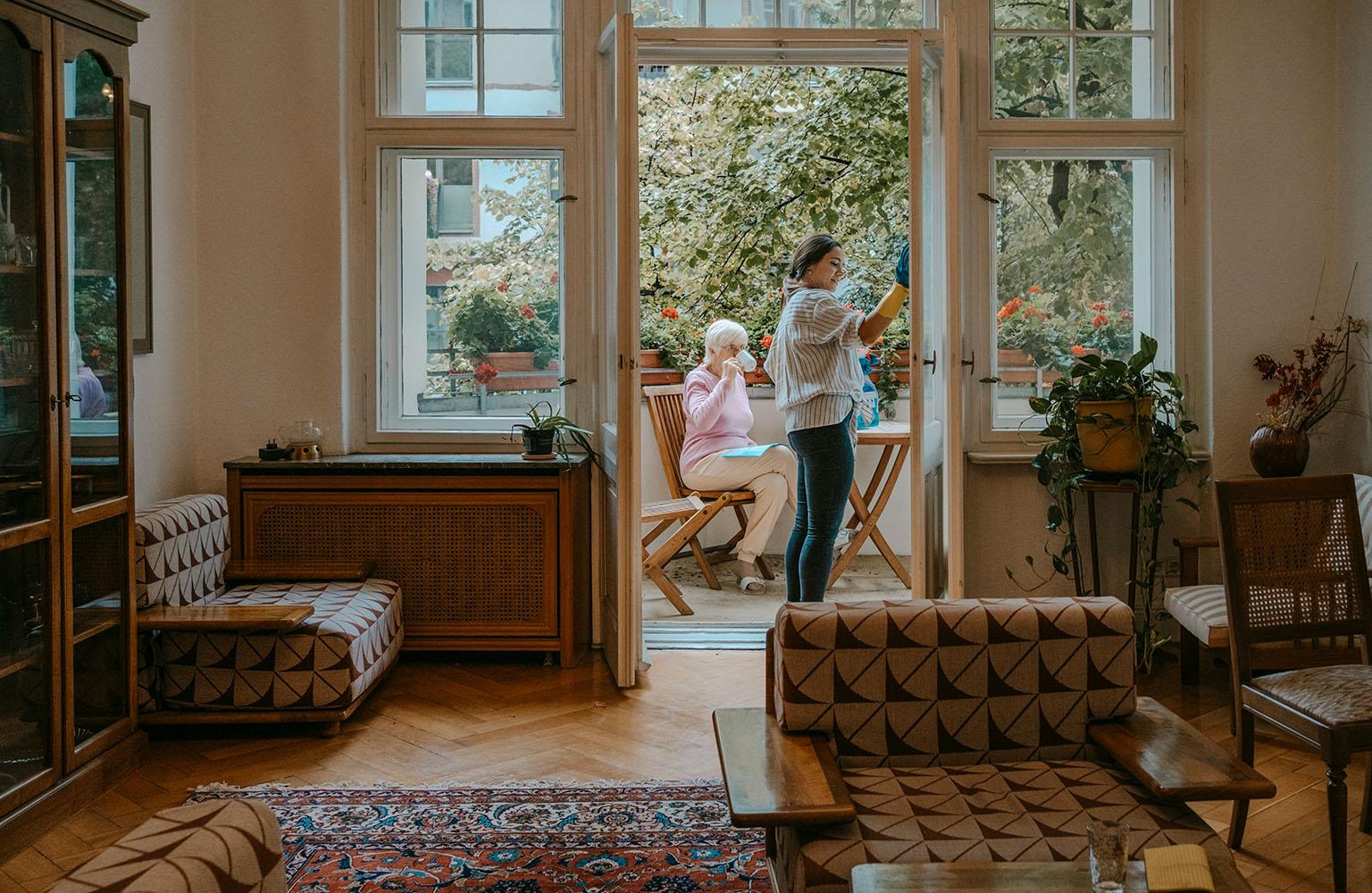 Ältere Dame trinkt auf dem Balkon Kaffee während eine andere Frau Fenster putzt