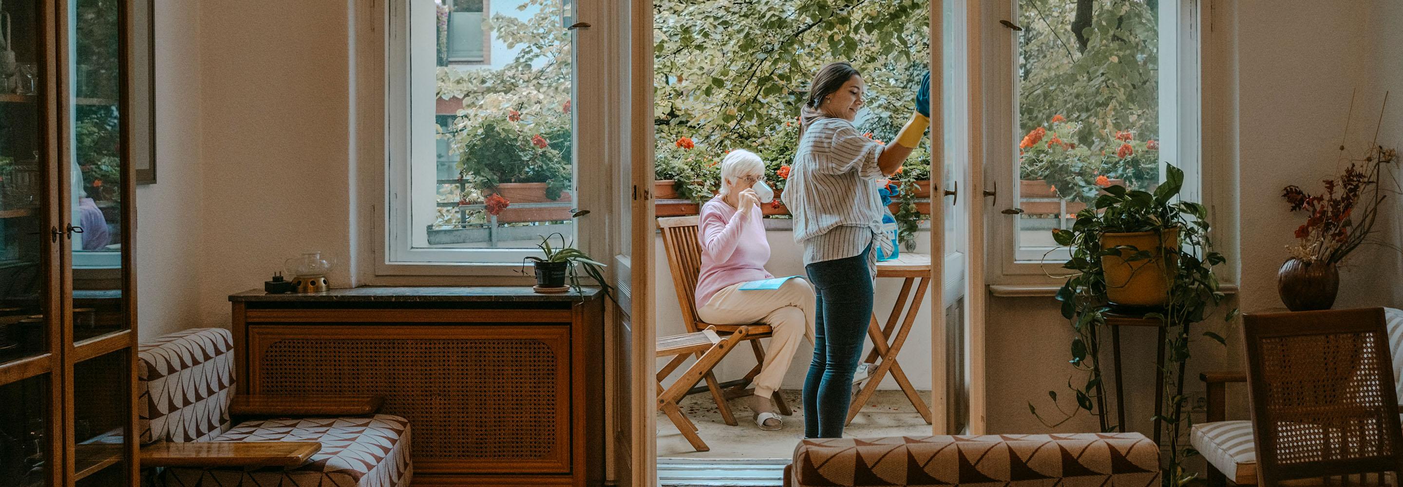 Ältere Dame trinkt auf dem Balkon Kaffee während eine andere Frau Fenster putzt