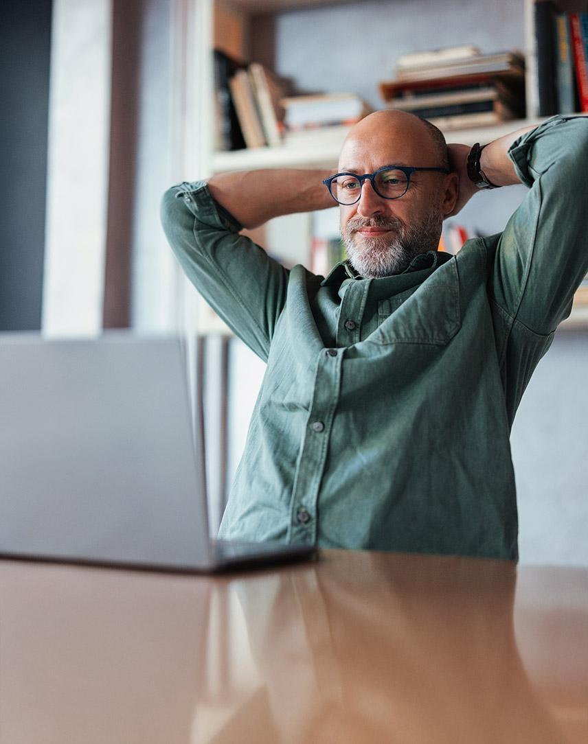 Mann schaut in seinen Laptop und verschränkt zufrieden seine Hände hinter dem Kopf