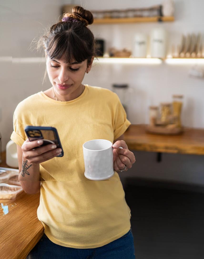 Frau mit Kaffeetasse in der Hand schaut in der Küche auf Smartphone