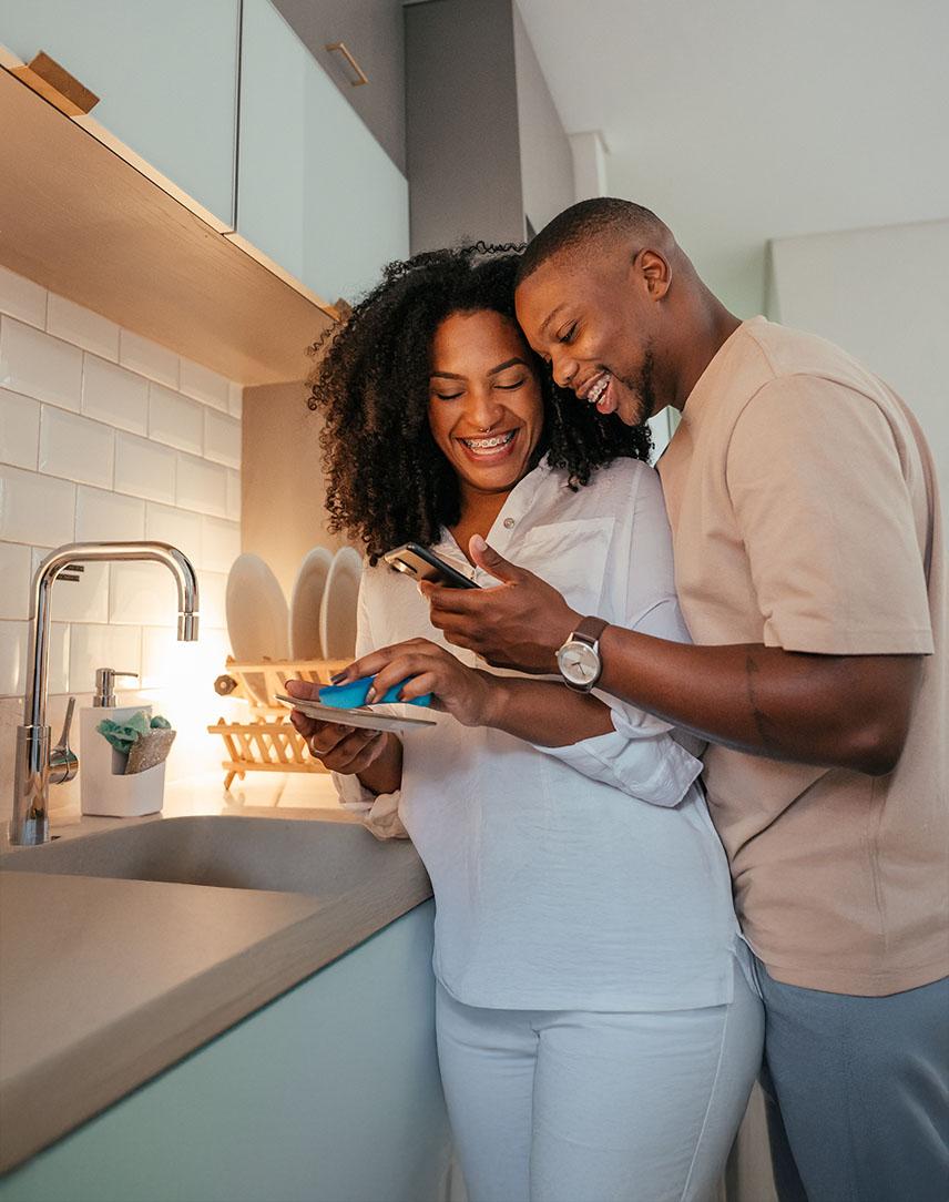 Mann und Frau stehen in der Küche und schauen lachend auf ein Smartphone