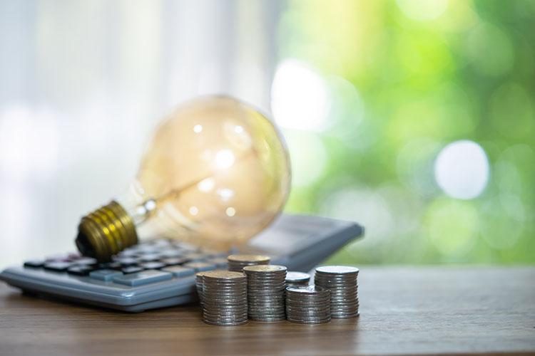 Glühbirne auf Taschenrechner liegend, davor gestapeltes Hartgeld