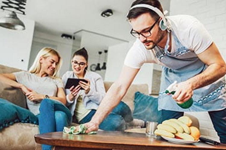 Zwei Freundinnen schauen auf ein Tablet, ein Mann putzt derweil den Couchtisch