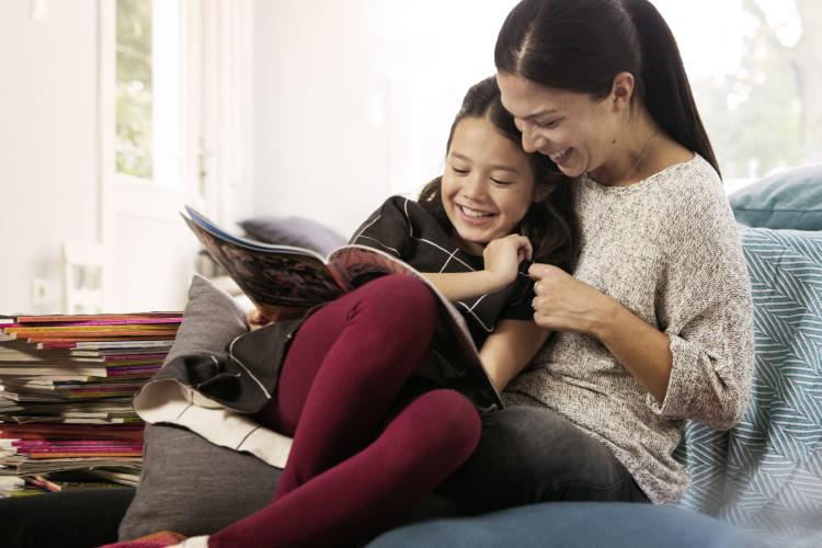Mutter sitzt mit ihrer Tochter auf der Couch und liest ihr aus Zeitschriften vor