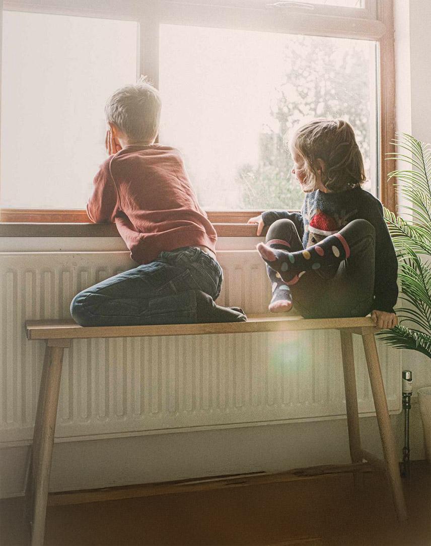 Zwei Kinder sitzen auf einer Bank vor der Heizung und schauen aus dem Fenster