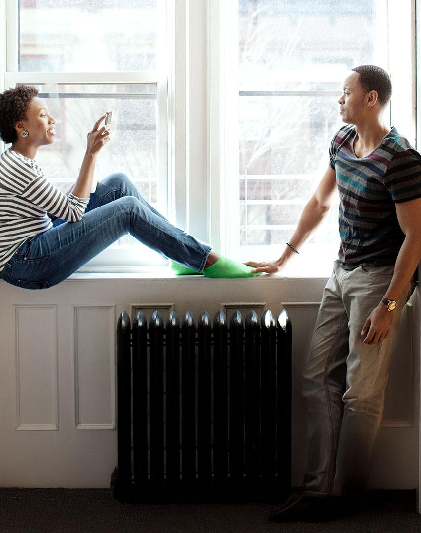Frau sitzt auf der Fensterbank über einer Heizung und unterhält sich mit Mann