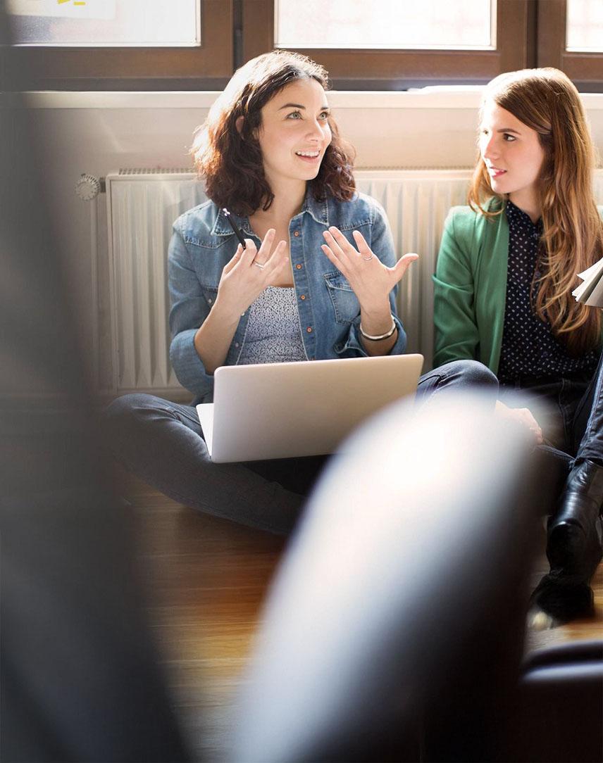 Zwei Frauen sitzen mit Laptop und Unterlagen auf dem Boden vor einer Heizung