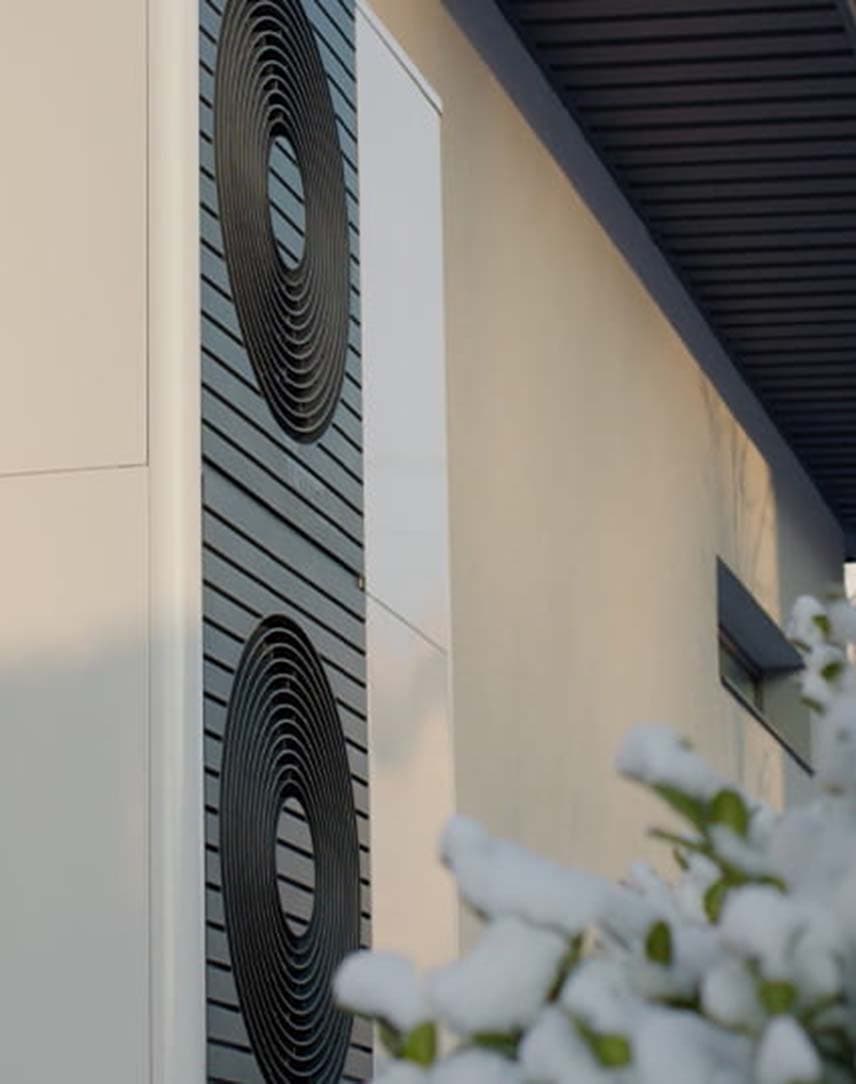 Wärmepumpe an einer Hauswand, davor ein eingeschneiter Busch