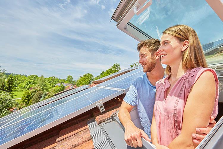 Mann und Frau schauen aus einem Dachfenster, daneben sind PV-Module sichtbar