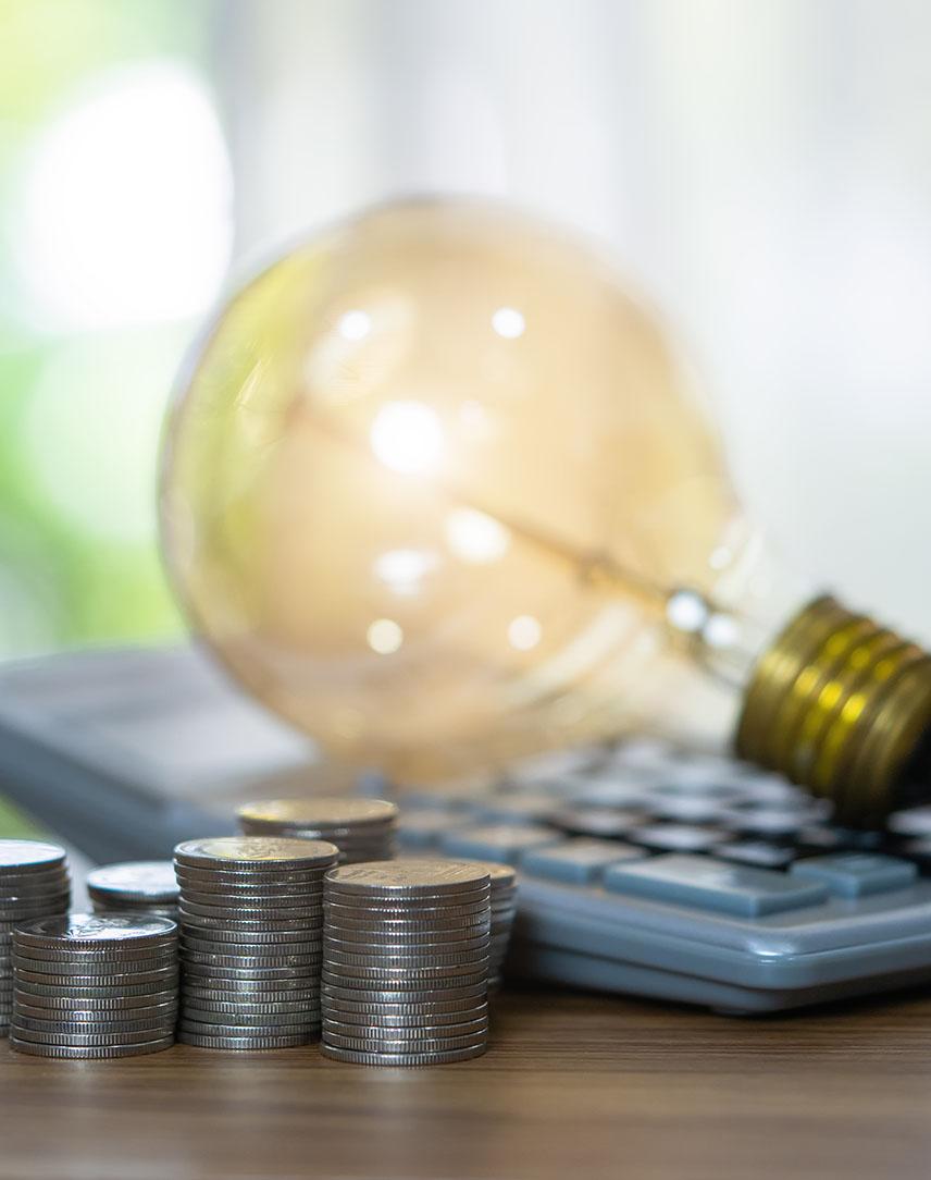 Glühbirne auf Taschenrechner liegend, davor gestapeltes Hartgeld